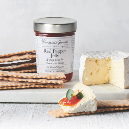 A white rustic tabletop with a white marble cheese board topped with a jar of Bonnie's red pepper jelly, flatbread crackers, a small wheel of soft cheese, and a cracker topped with cheese and red pepper jelly
