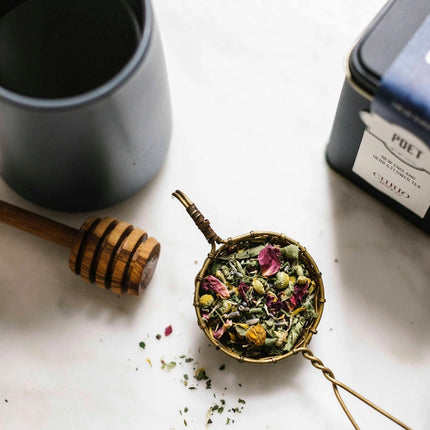 a tea infuser filled with a colorful floral tea next to the tea tin, a storage pot, and a honey server