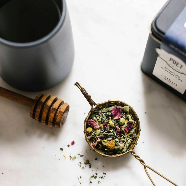 a tea infuser filled with a colorful floral tea next to the tea tin, a storage pot, and a honey server