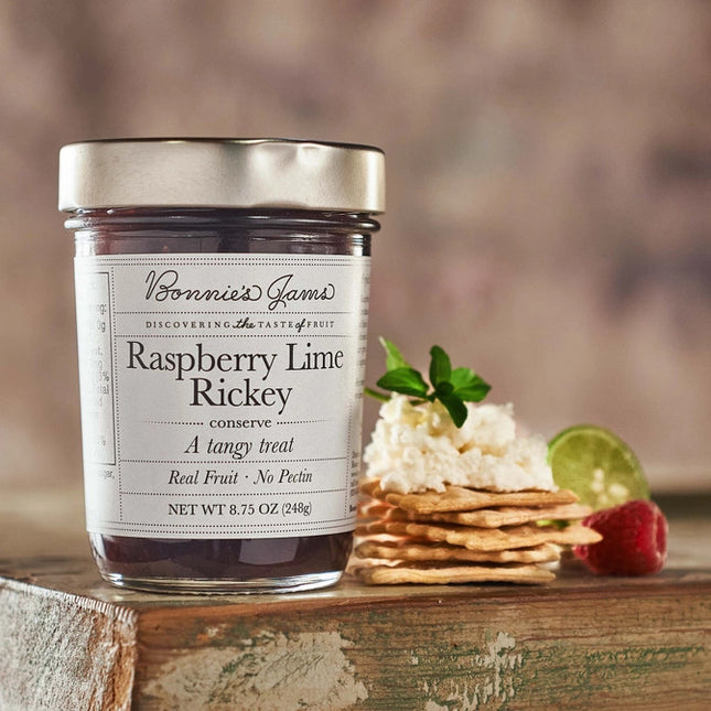 Bonnie's Raspberry Lime Rickey jam jar with an elegant white label and silver jar top on a table next to a stack of crackers topped with a soft cheese and herbs