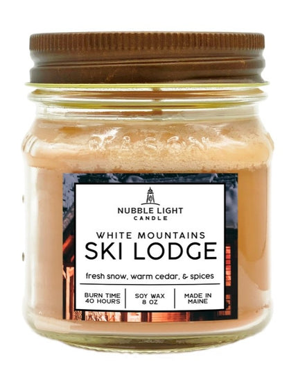 a candle in a clear glass mason jar with tan wax and a label that reads "White Mountains Ski Lodge" against a white background