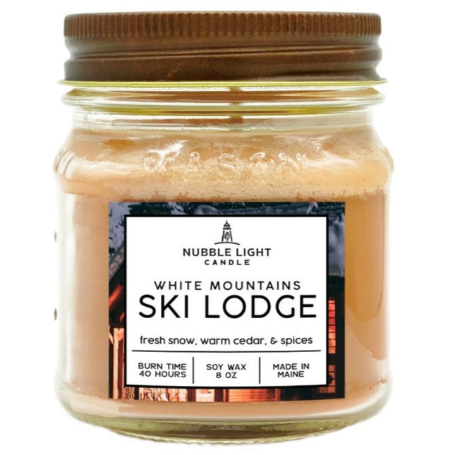 a candle in a clear glass mason jar with tan wax and a label that reads "White Mountains Ski Lodge" against a white background