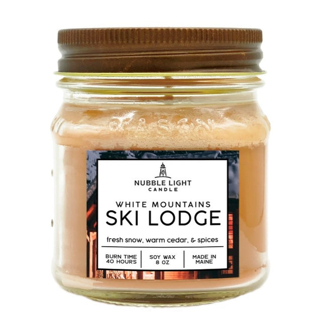 a candle in a clear glass mason jar with tan wax and a label that reads "White Mountains Ski Lodge" against a white background