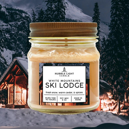 a candle in a clear glass mason jar with tan wax and a label that reads "White Mountains Ski Lodge" against an image of a snowy mountain with a ski cabin