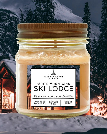a candle in a clear glass mason jar with tan wax and a label that reads "White Mountains Ski Lodge" against an image of a snowy mountain with a ski cabin