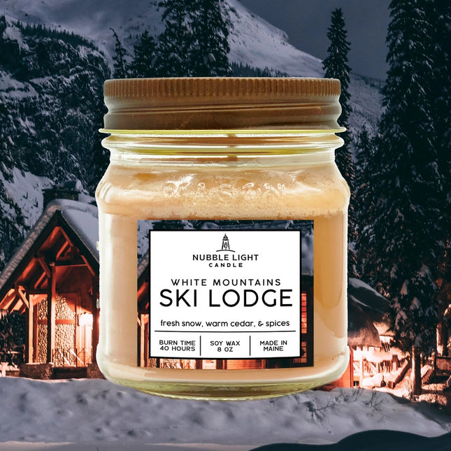 a candle in a clear glass mason jar with tan wax and a label that reads "White Mountains Ski Lodge" against an image of a snowy mountain with a ski cabin