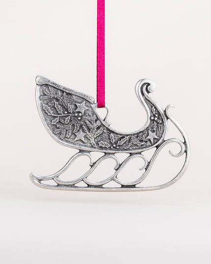 the back of the ornament with hand engraved maker's mark "danforth pewter 1992" - Handcrafted pewter sleigh ornament hung from a red satin ribbon, the sleigh is adorned with pine bough, holly, and star details
