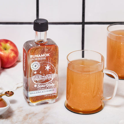 snow globe sparkle syrup on a countertop next to apples and clear mugs showing a sparkly cider drink