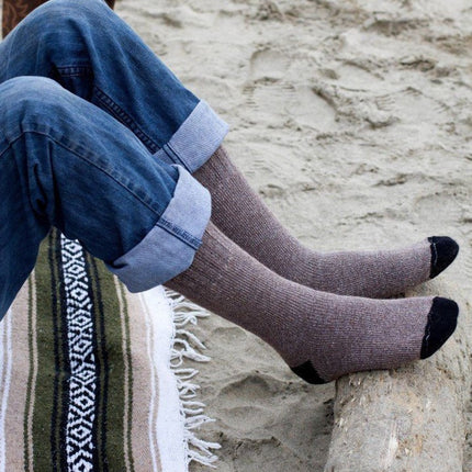 a pair of legs wearing rolled up jeans and cocoa brown alpaca socks resting their feet on a log with beach sand visible in the background