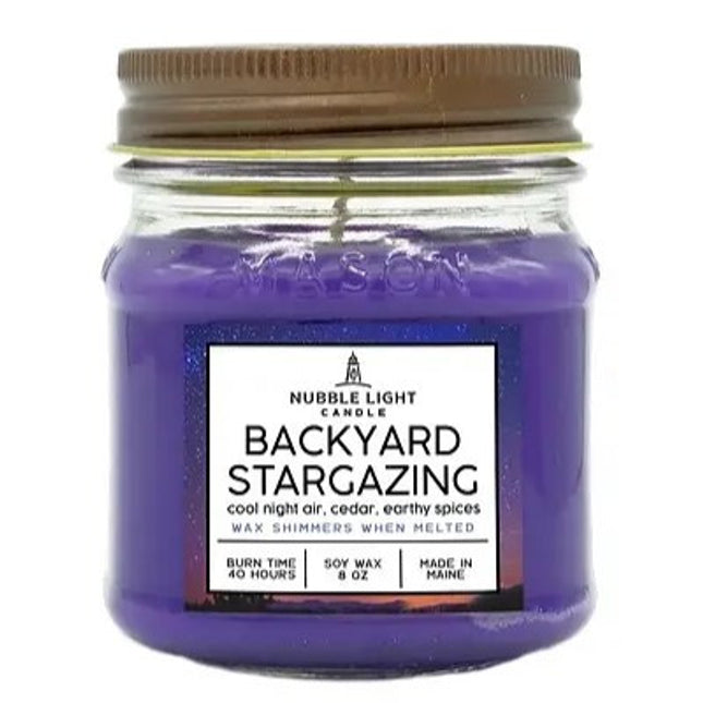 a candle in a clear glass mason jar with purple wax and a label that reads "Backyard Stargazing" against a white background