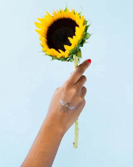 an outstretched hand holding a sunflower with a honey bee temporary tattoo on the model's wrist against a sky blue background
