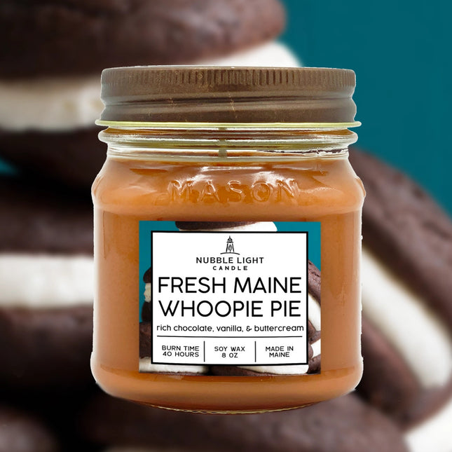 a candle in a clear glass mason jar with light brown wax and a label that reads "Maine Whoopie Pie" against a soft image of a pile of whoopie pies