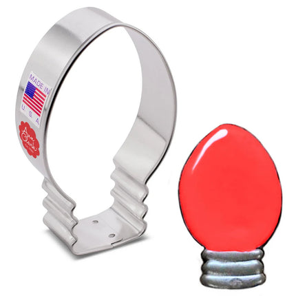 a silver, tin cookie cutter in the shape of a Christmas light bulb - oblong shape with a soft point at the top, next to a fully decorated cookie of a Christmas light bulb with red icing on the bulb and sliver icing on the base of the bulb.